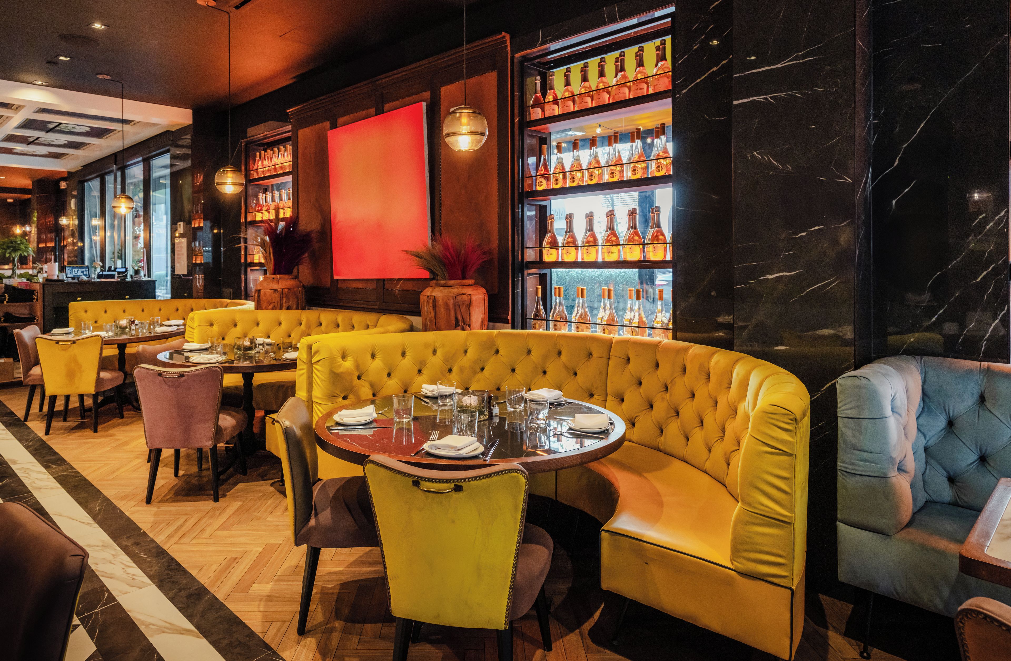 A restaurant interior with yellow booths, round tables, and a wine display.