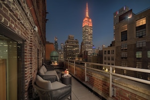 A rooftop terrace with wicker furniture, a small table with candles, and a view of the Empire State Building at dusk.