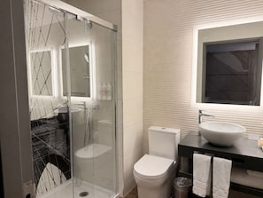 A modern bathroom with a glass-enclosed shower, a white toilet, a sink with a large mirror above it, and a towel rack with two towels. - NH Collection New York Madison Avenue (New York)