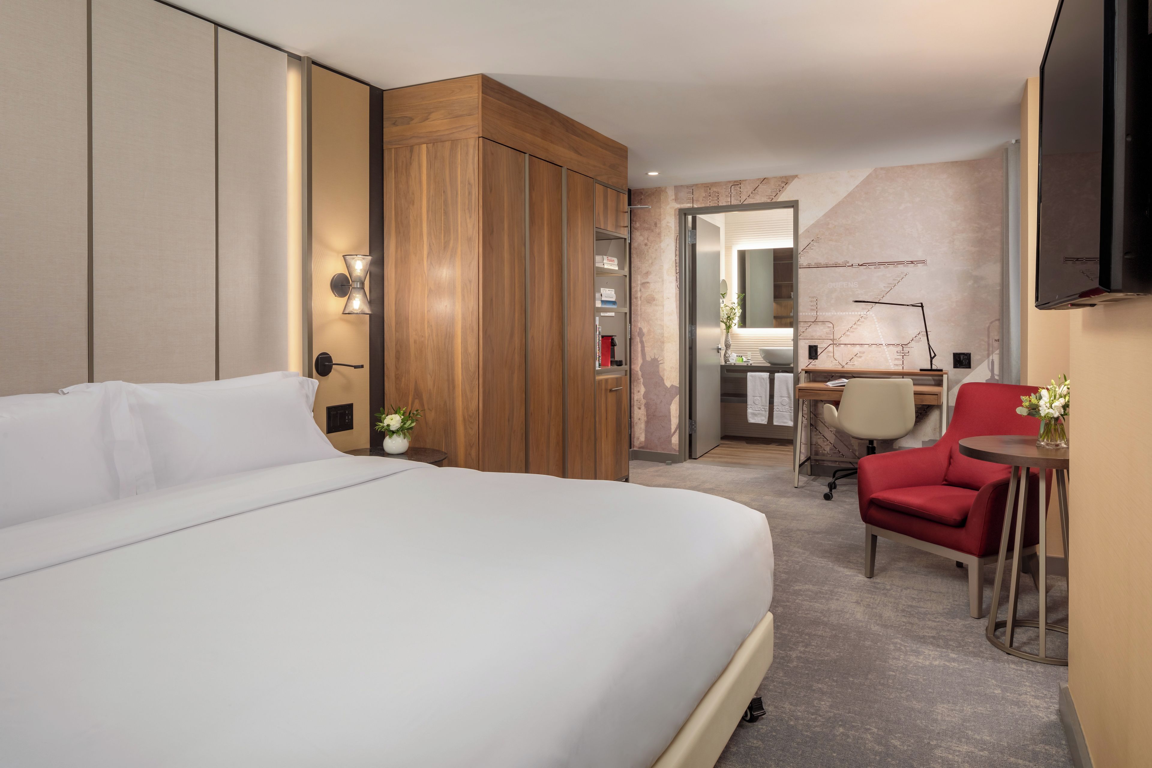 Ein Hotelzimmer mit einem großen Bett, einem Schreibtisch mit Stuhl, einem roten Sessel und einem kleinen Tisch mit Blumen.