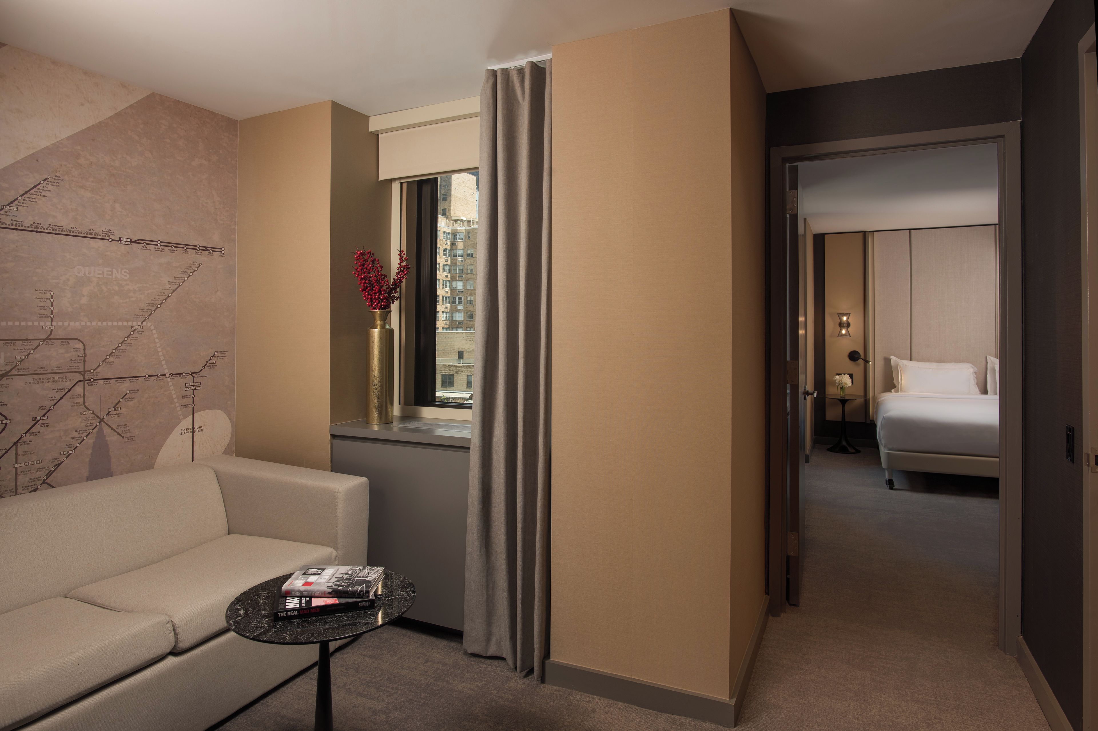 A hotel room with a sofa, a small table with books, a window with curtains, and a view of buildings outside.