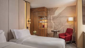 A hotel room with two beds, a wooden cabinet with shelves, a small table with a lamp, and a red chair.