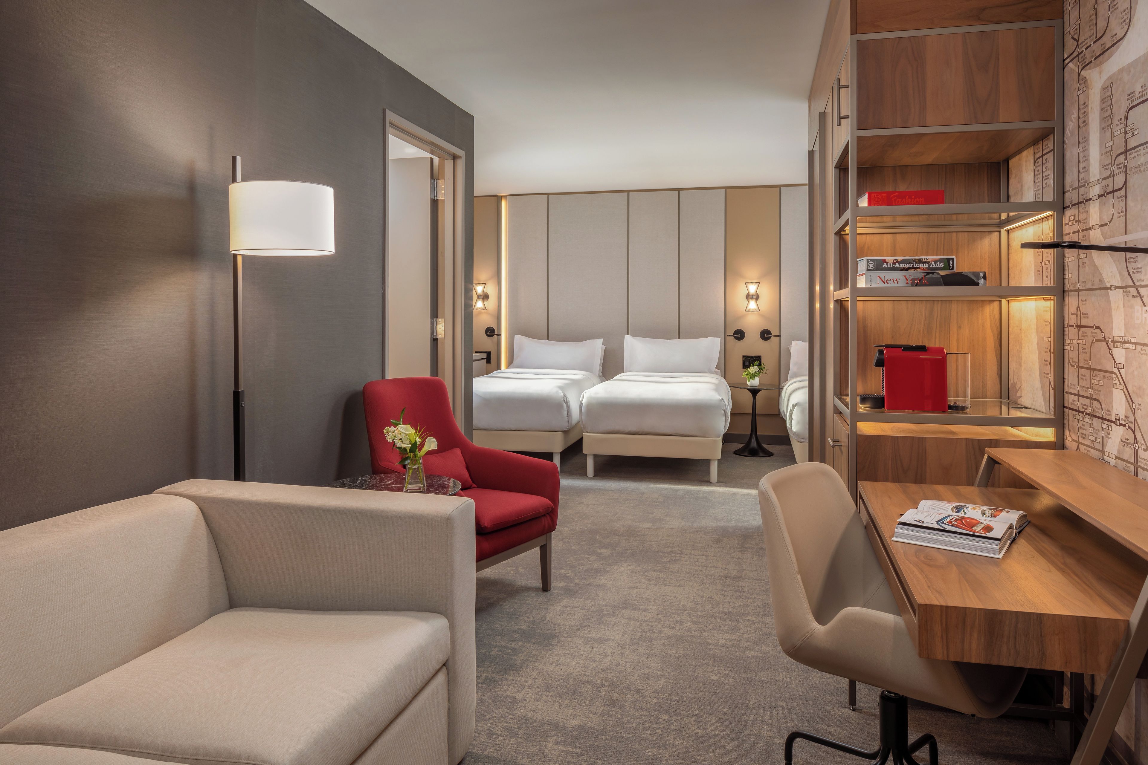 A hotel room with a sofa, a red armchair, a desk, a bookshelf, and a bed with two pillows.