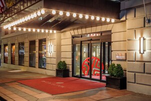 Entrada de un hotel con alfombra roja, un letrero iluminado con el número '6' y una puerta de cristal.