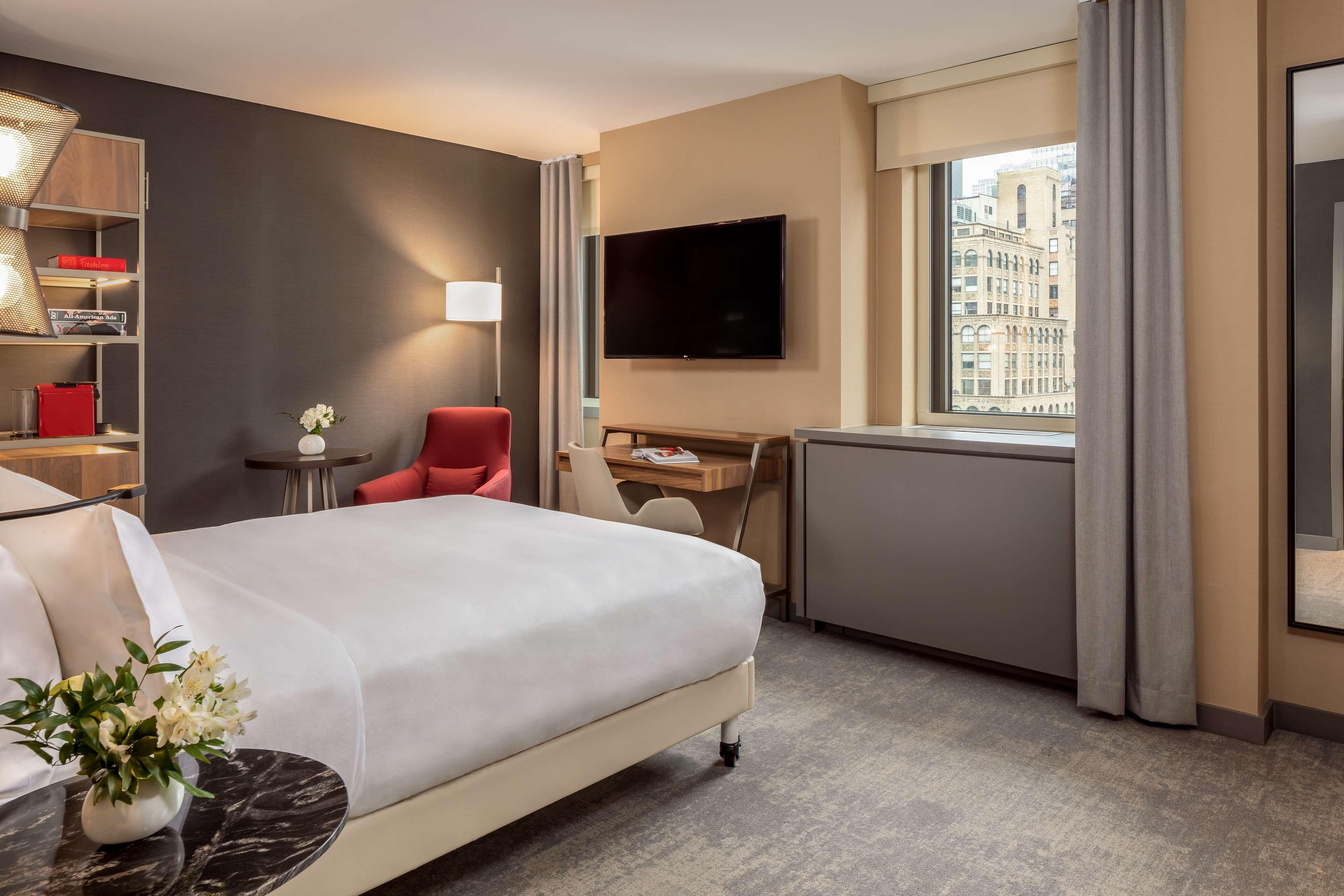 Une chambre d’hôtel équipée d’un lit, d’un bureau, d’une chaise, d’une télévision et d’une fenêtre donnant sur la ville.
