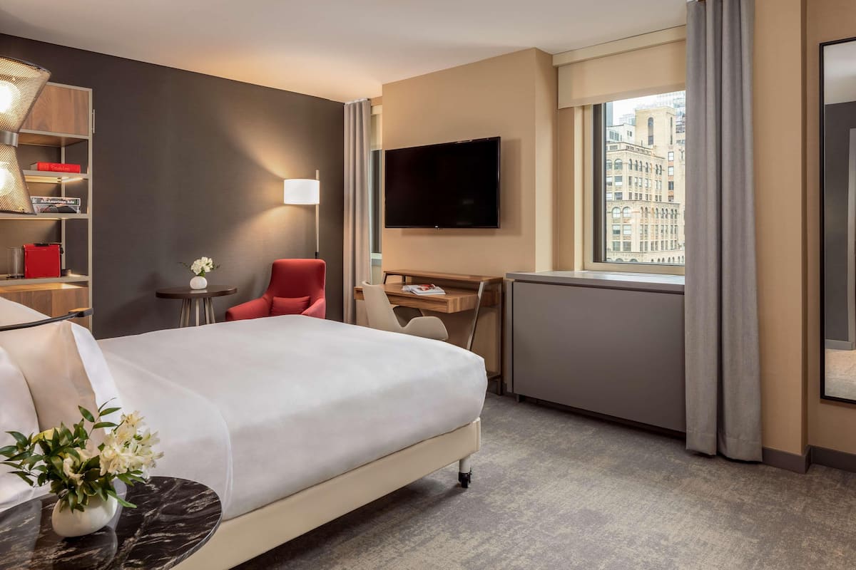 Ein Hotelzimmer mit Bett, Schreibtisch, Sessel, Fernseher und einem Fenster mit Blick auf die Stadt.