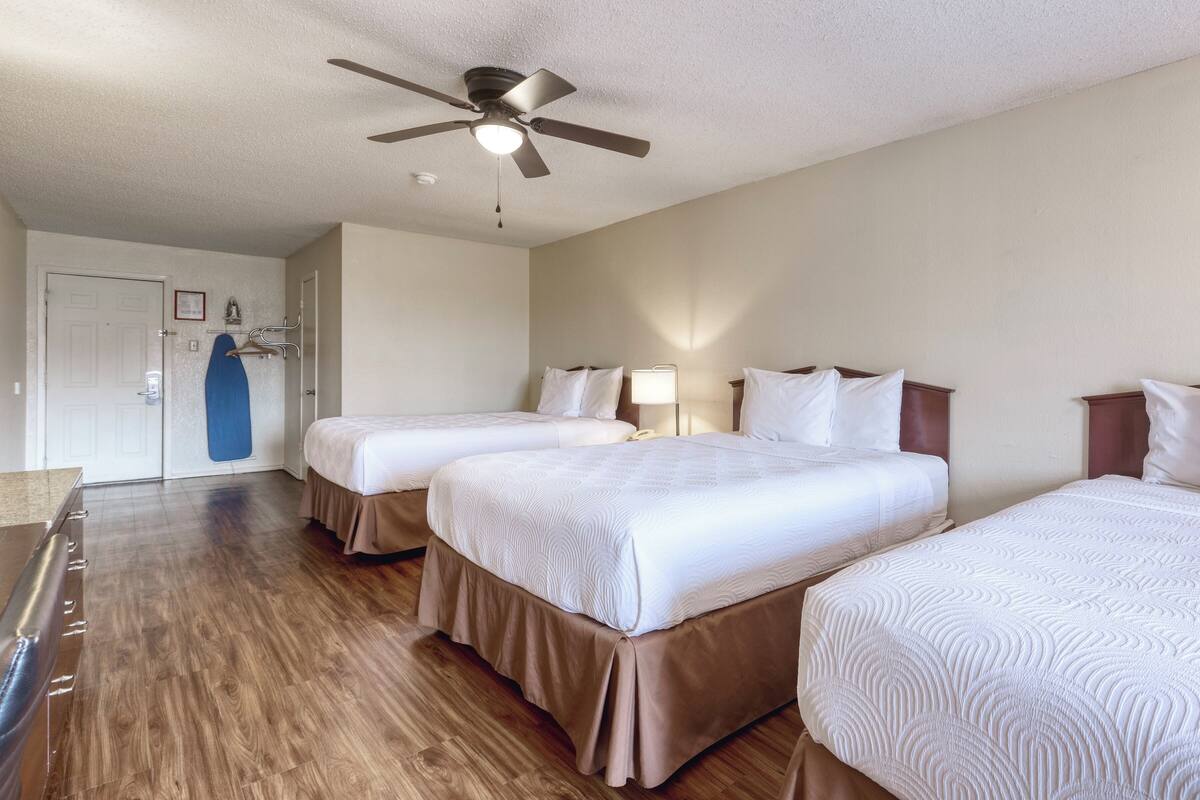 A hotel room with two beds, a ceiling fan, a door, and a blue surfboard on the wall.