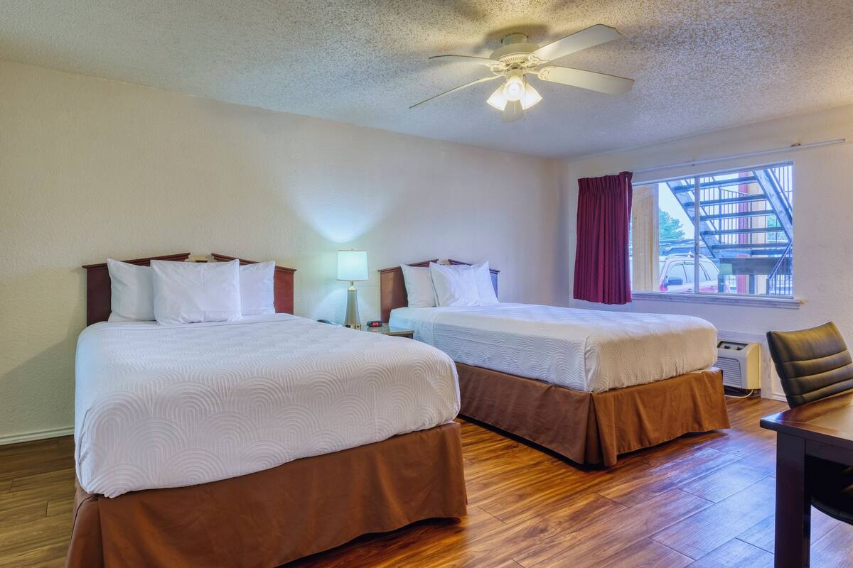A hotel room with two beds, a ceiling fan, a window with a view of a building, and a table with a chair.