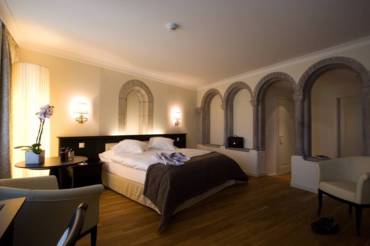 A hotel room with a large bed, a sitting area with a chair, a small table with a lamp, and a potted plant.