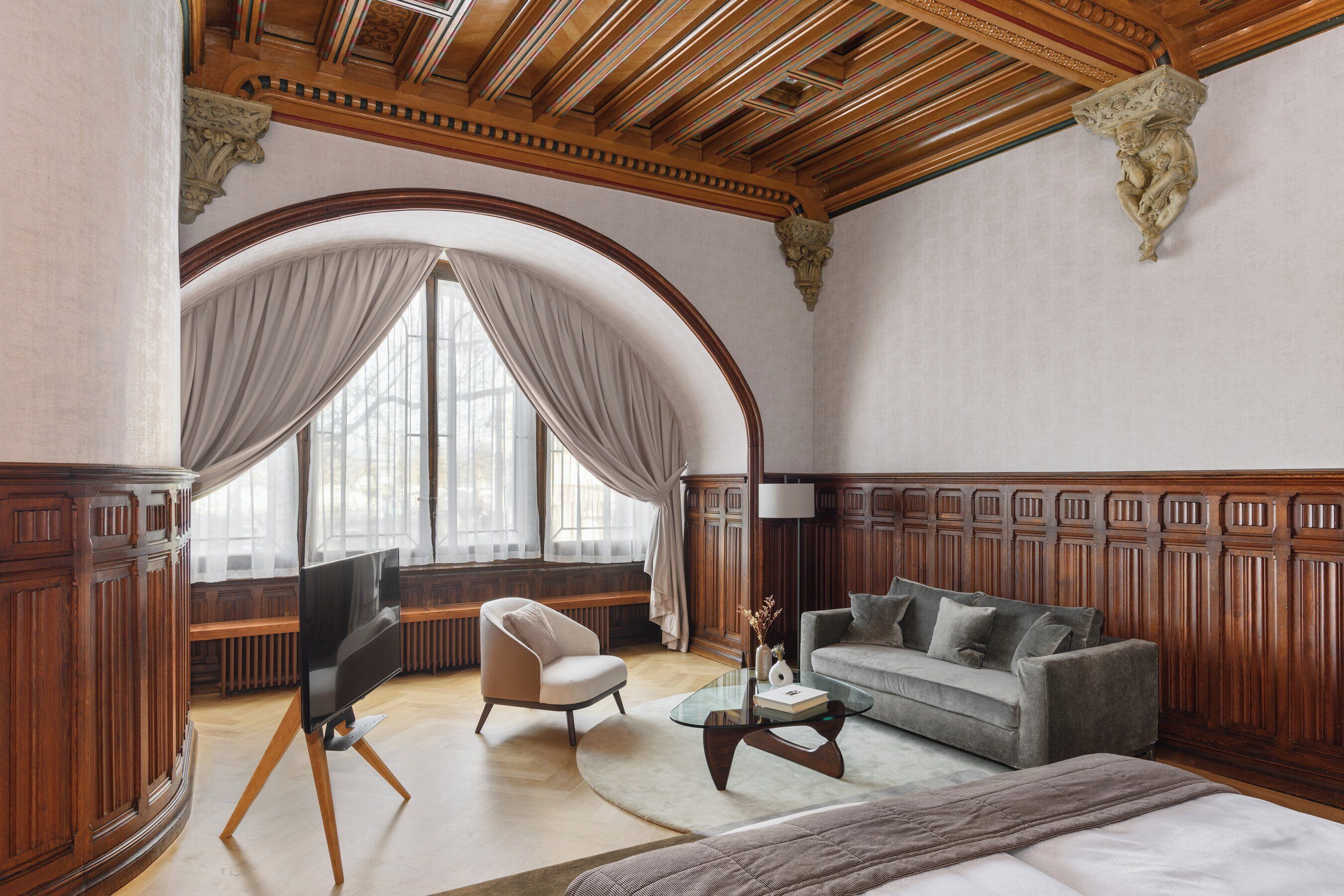 Una habitación con un amplio ventanal arqueado, un sofá, una silla, una mesa de centro y un televisor sobre un mueble.