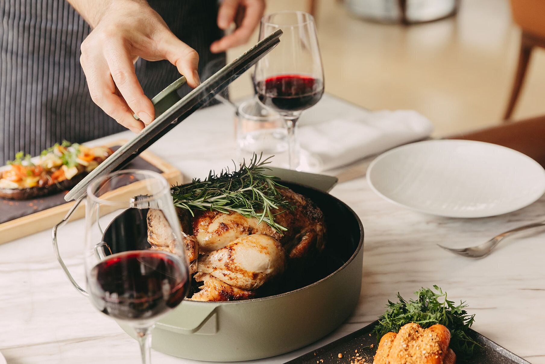 A dining table with a roasted chicken dish, a glass of red wine, and a pair of chopsticks.
