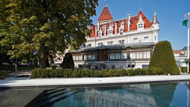 A large, historic building with a red-tiled roof and multiple towers, adjacent to a swimming pool with lounge chairs. - Château d'Ouchy (Lausanne)
