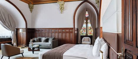 A bedroom with a large bed, a sofa, a chair, a glass coffee table, and a stained glass window.