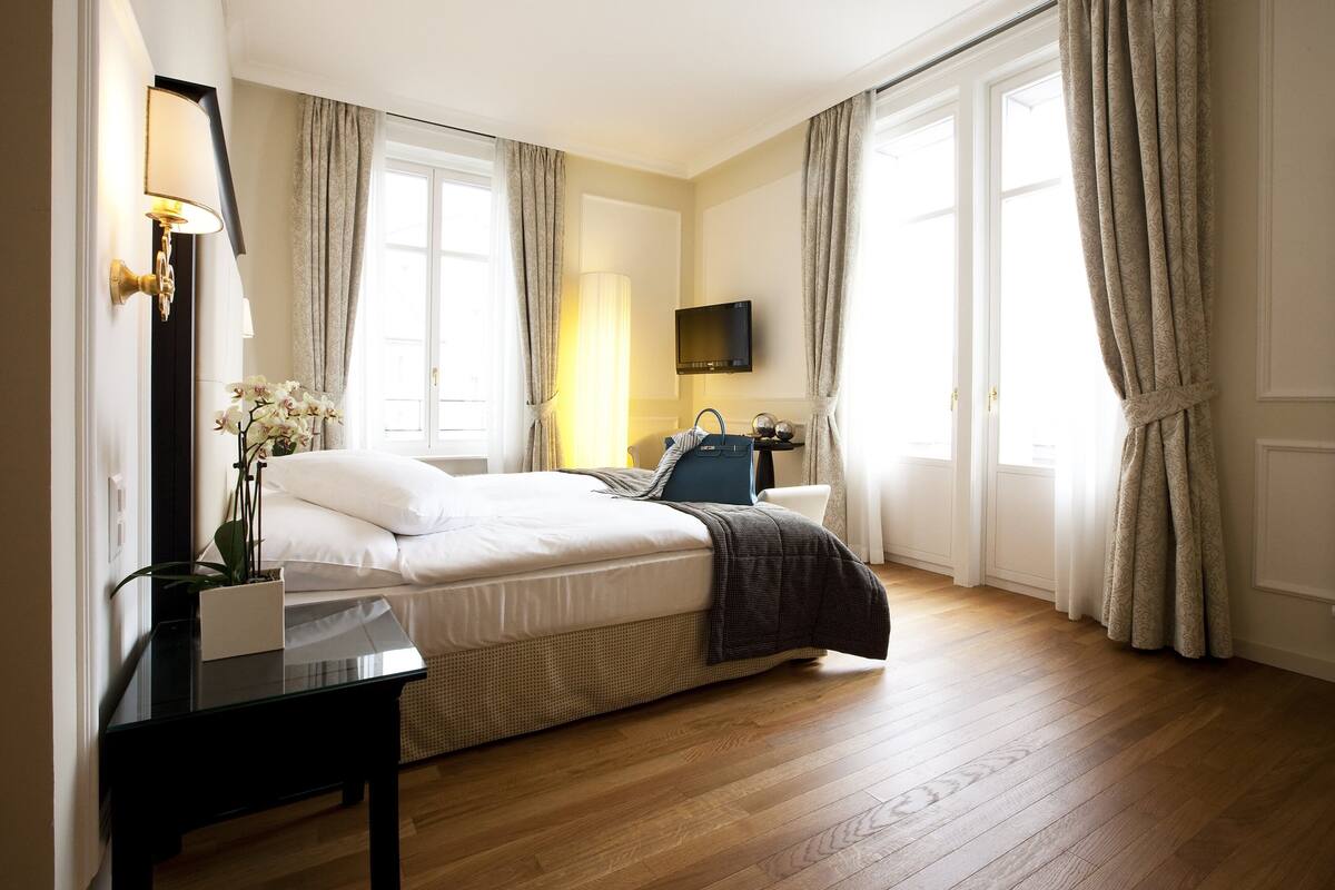 A hotel room with a bed, bedside table, lamp, TV, and curtains.