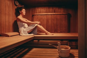 A person sitting in a wooden sauna with benches and a wooden bucket.