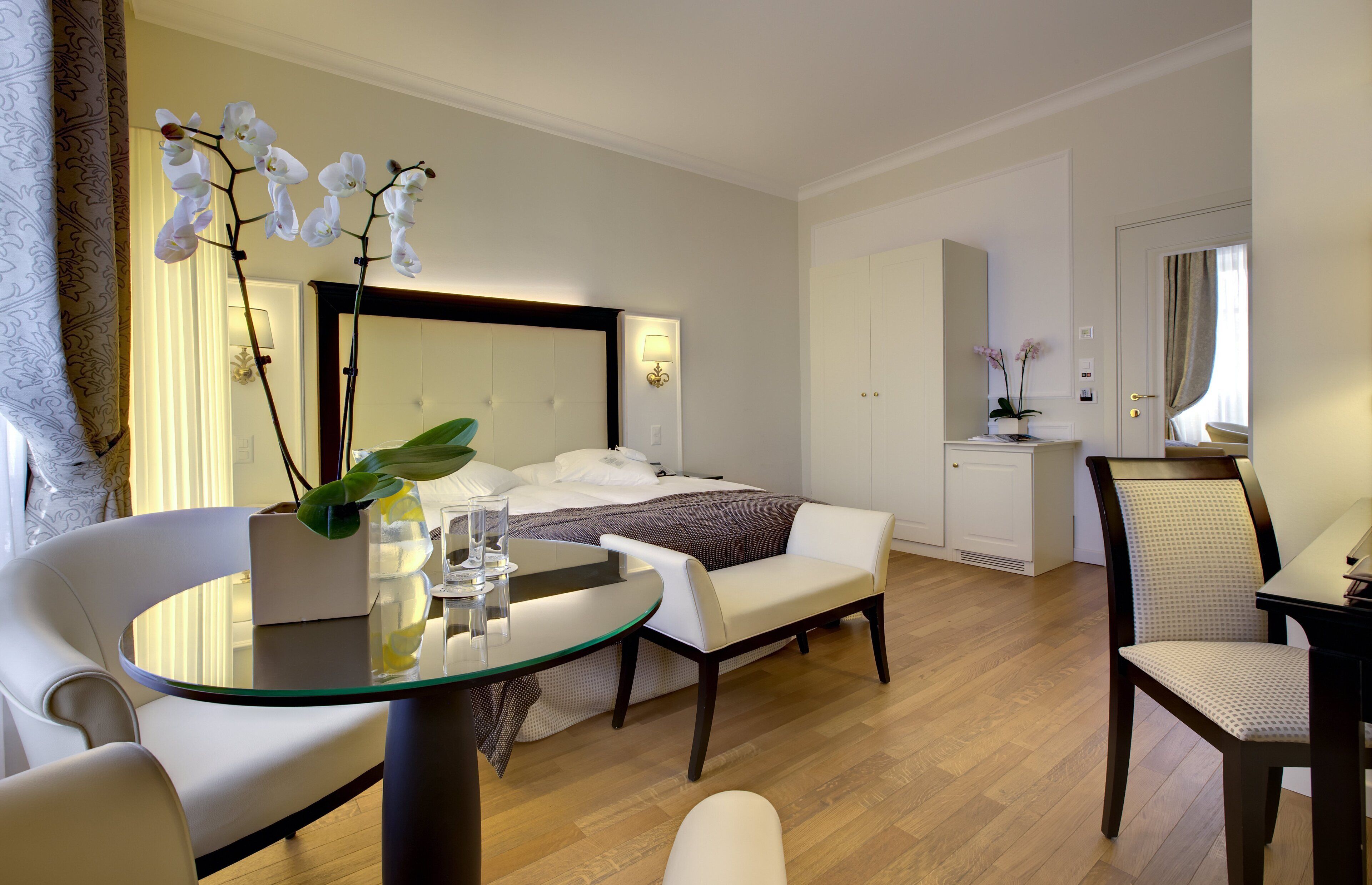 A modern hotel room with a bed, a sitting area with a glass table, a chair, and a desk.