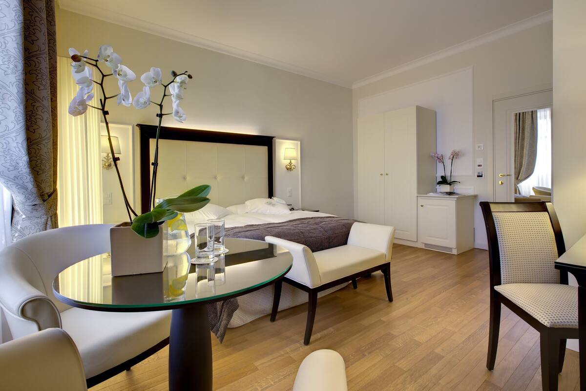 A modern hotel room with a bed, a sitting area with a glass table, a chair, and a desk.