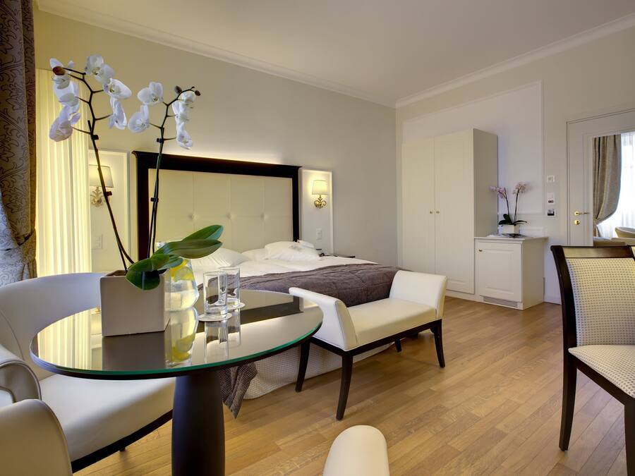A modern hotel room with a bed, a sitting area with a glass table, a chair, and a desk.