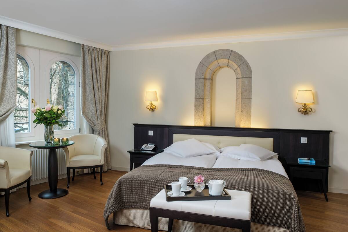A hotel room with a large bed, two bedside tables, a round table with chairs, and a window with curtains.