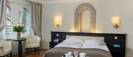 Une chambre d’hôtel avec un grand lit, deux tables de chevet, une petite table ornée d’un vase et de bougies, une chaise et une fenêtre avec des rideaux.