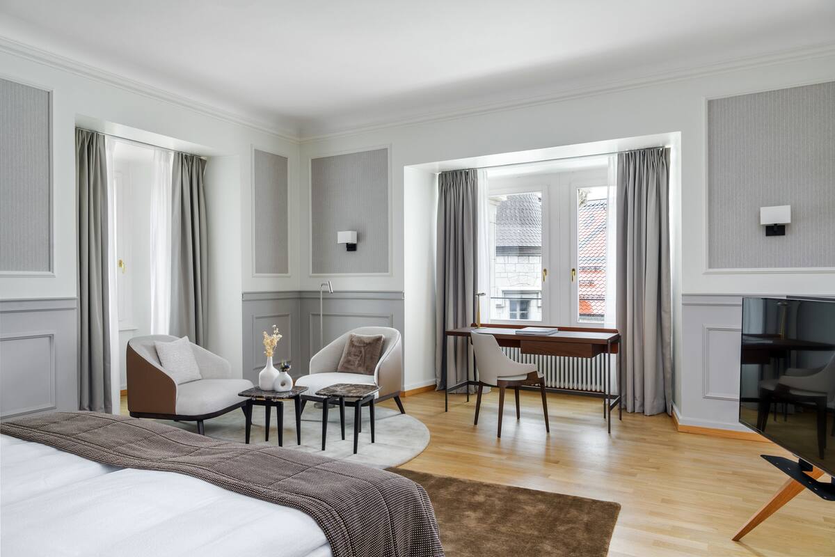 A modern hotel room with a large bed, two armchairs, a small table with a vase, a desk with a chair, a TV, and a view of a building through large windows.