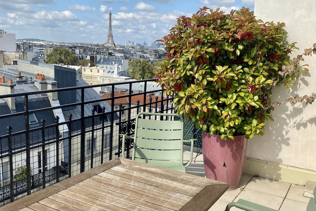 Un balcón con vista a la Torre Eiffel, una mesa de madera y sillas verdes.