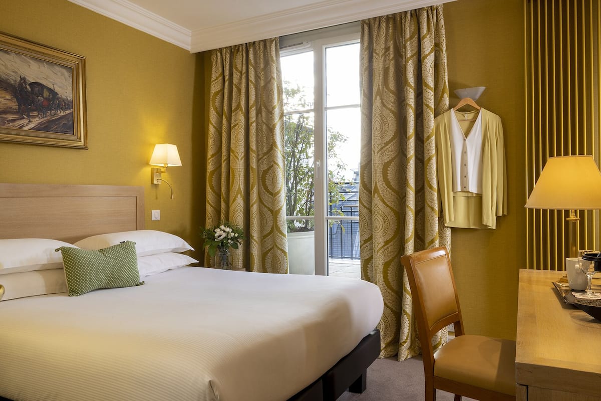 Habitación de hotel con una cama grande, una silla, una mesa con lámpara y vistas al exterior a trav és de una ventana con cortinas.