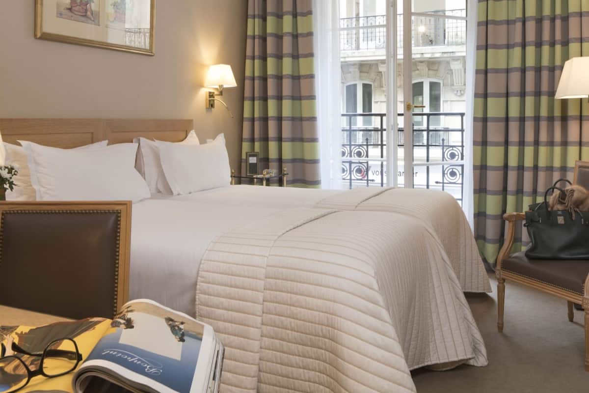 Habitación de hotel con cama doble, mesita de noche con lámpara, una silla y vistas a un balcón con barandilla.