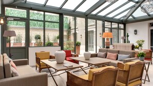 A spacious conservatory with a high ceiling, large windows, and a variety of seating arrangements including sofas, chairs, and tables. - Hôtel Le Littré (Paris)