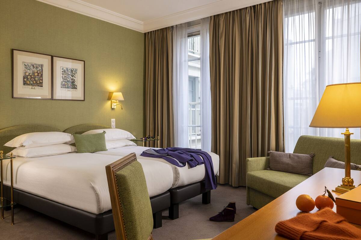 Habitación de hotel con cama, escritorio, silla, lámpara y ventana con cortinas.