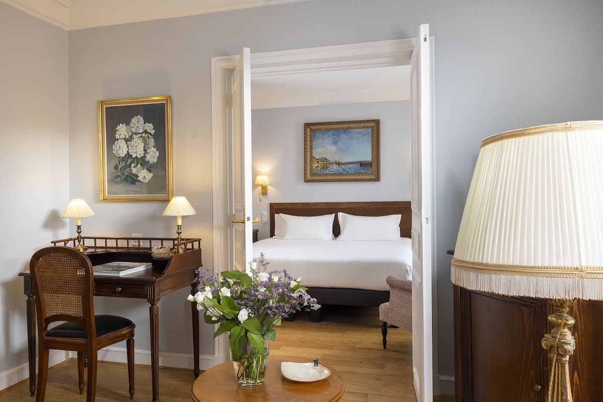 Una habitación de hotel con una cama, un escritorio con silla, un florero con flores y una lámpara.