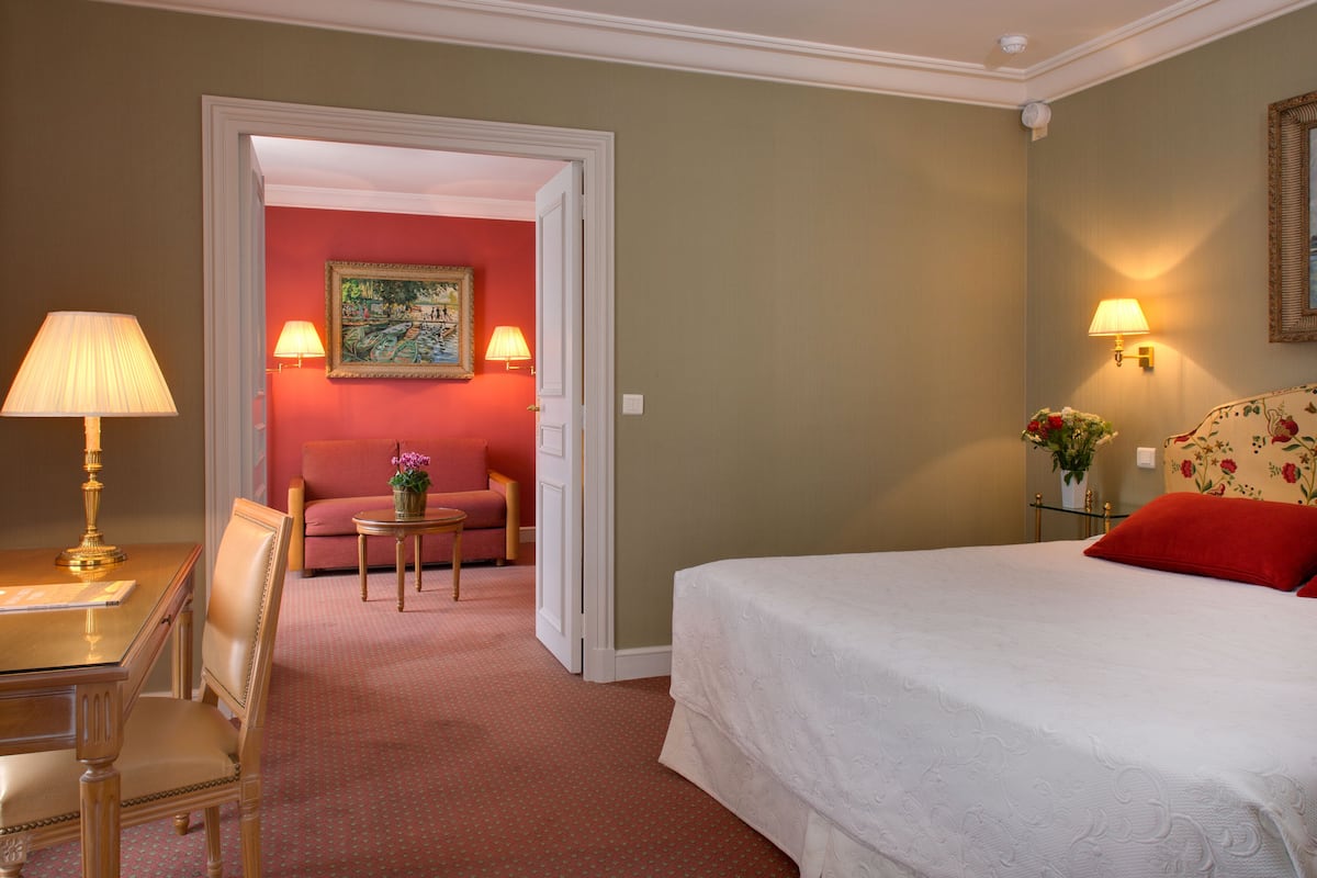 Una habitación de hotel con cama, escritorio con lámpara, silla, mesita con un florero y sofá.