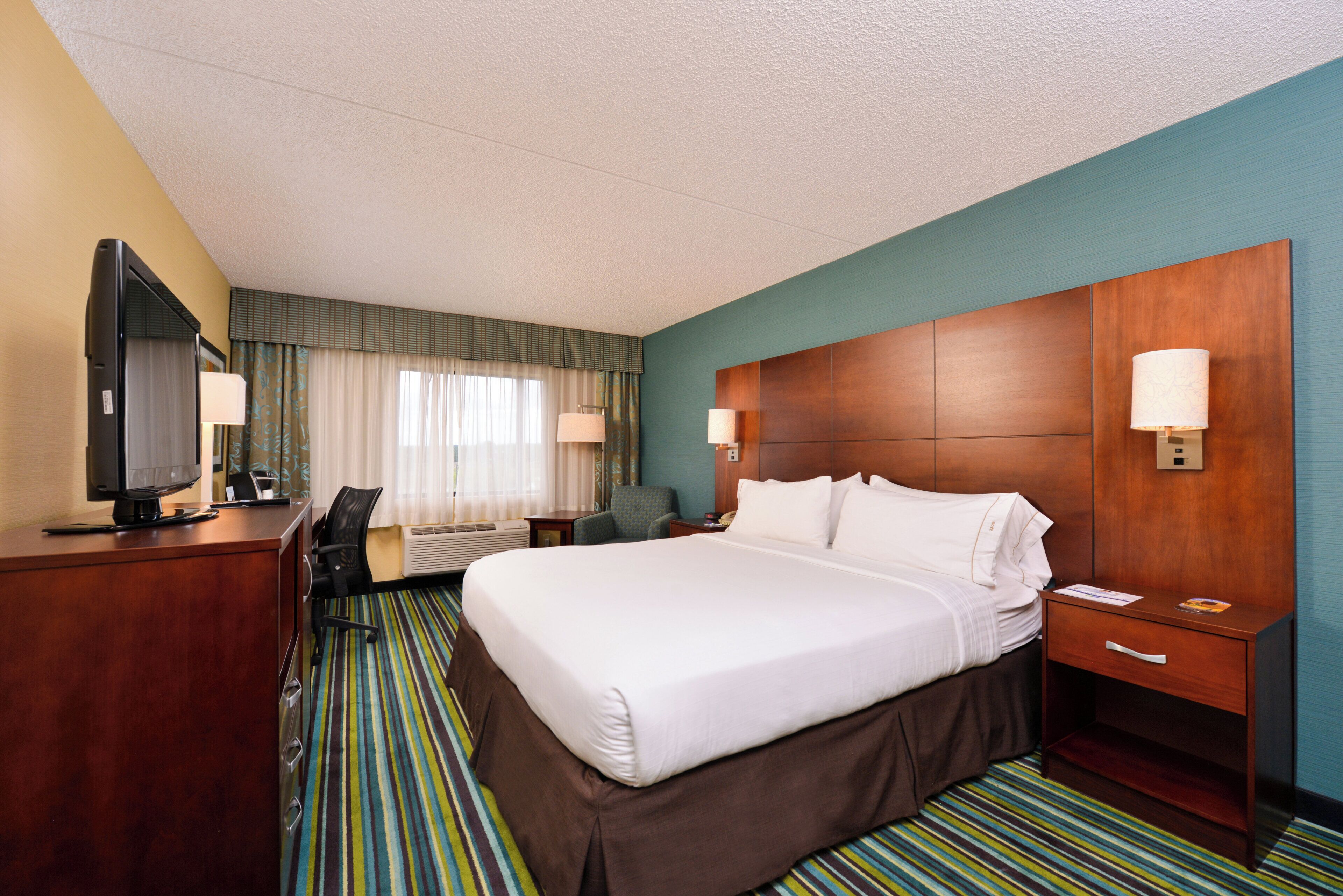 Quarto de hotel com uma cama grande, uma mesa de escritório com televisão, uma cadeira e uma mesa de cabeceira com abajur.