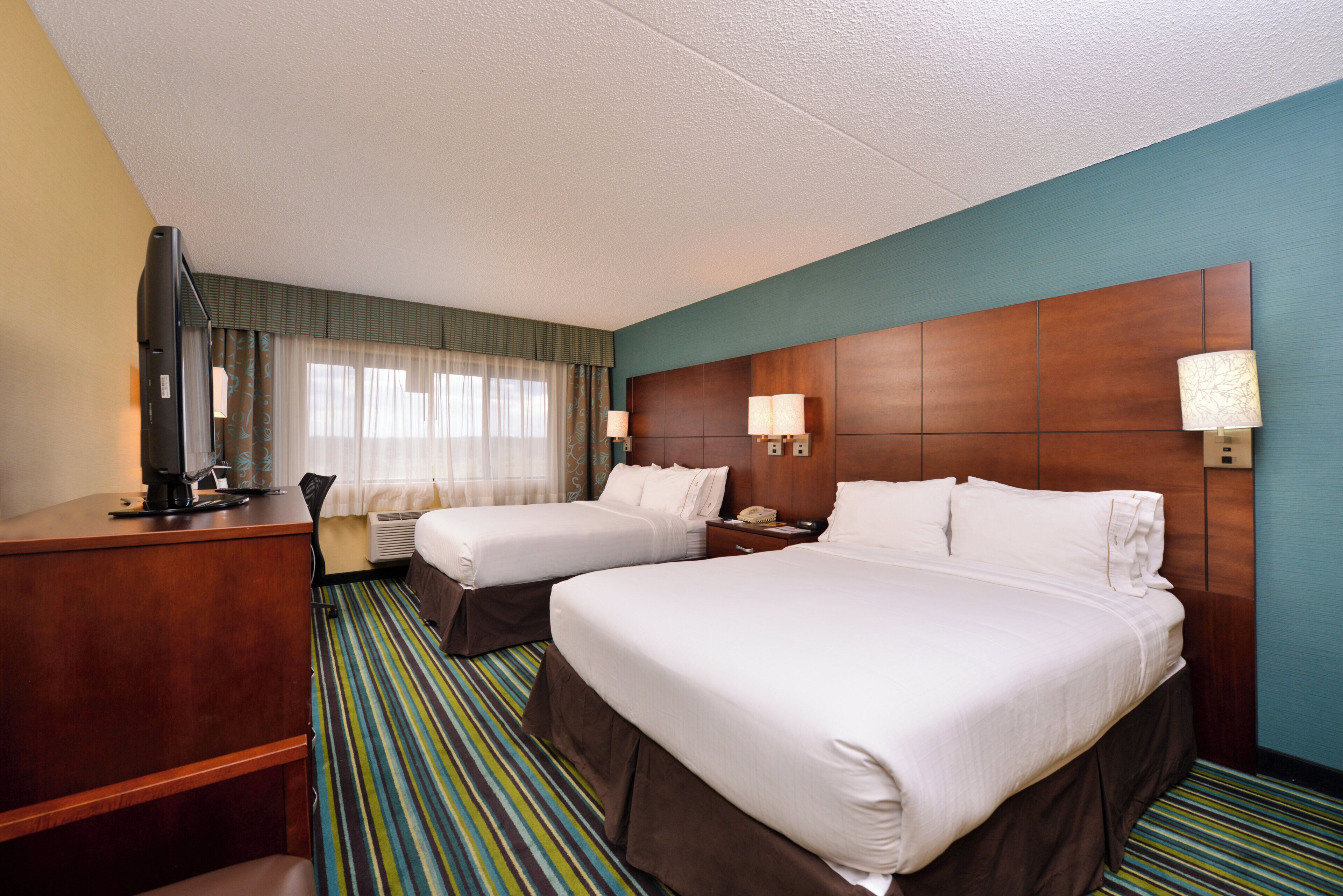 A hotel room with two beds, a desk, a chair, a lamp, and a window with curtains.