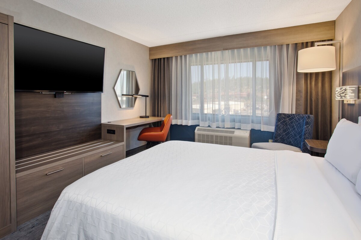 A hotel room with a large bed, a desk with a chair, a flat-screen TV, and a window with curtains.