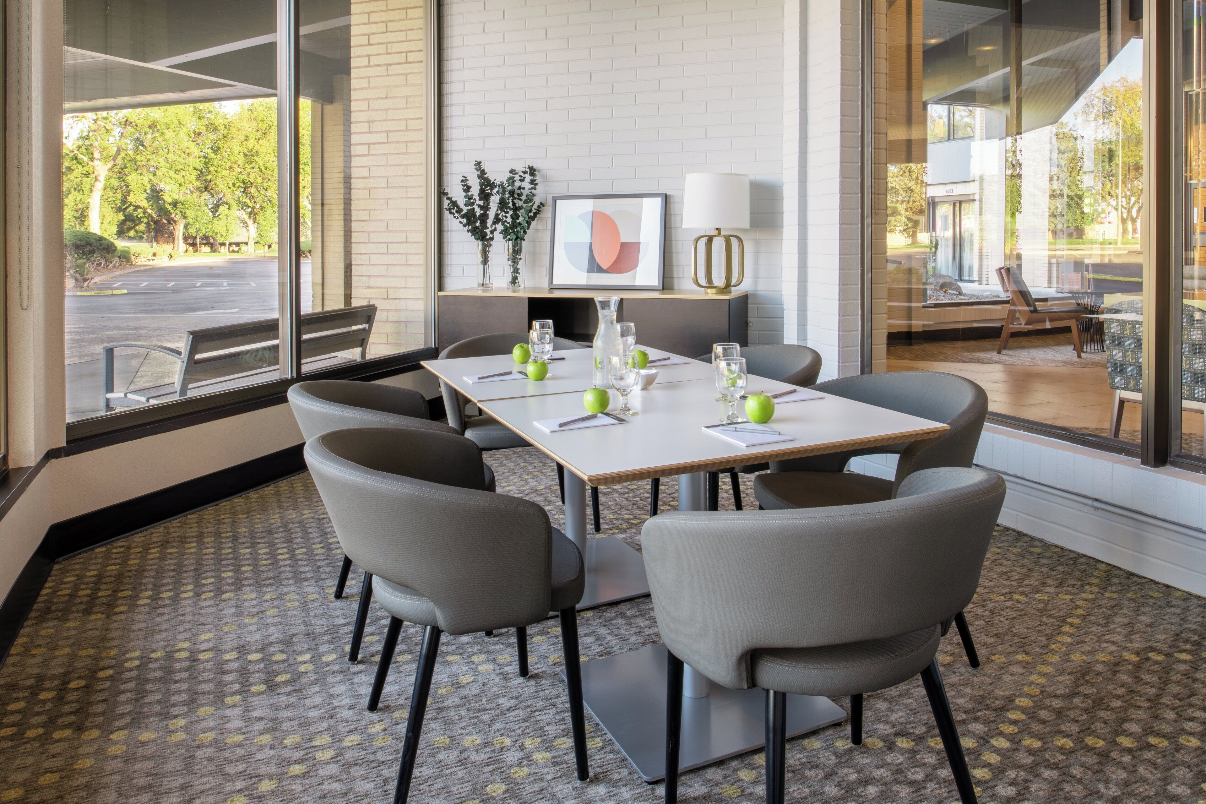 A conference room with a white table, grey chairs, and a view of the outdoors through large windows.