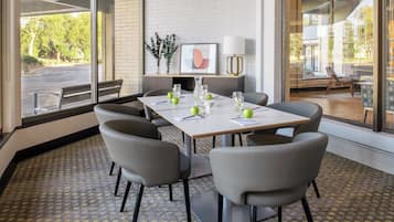 A conference room with a white table, grey chairs, and a view of the outdoors through large windows.