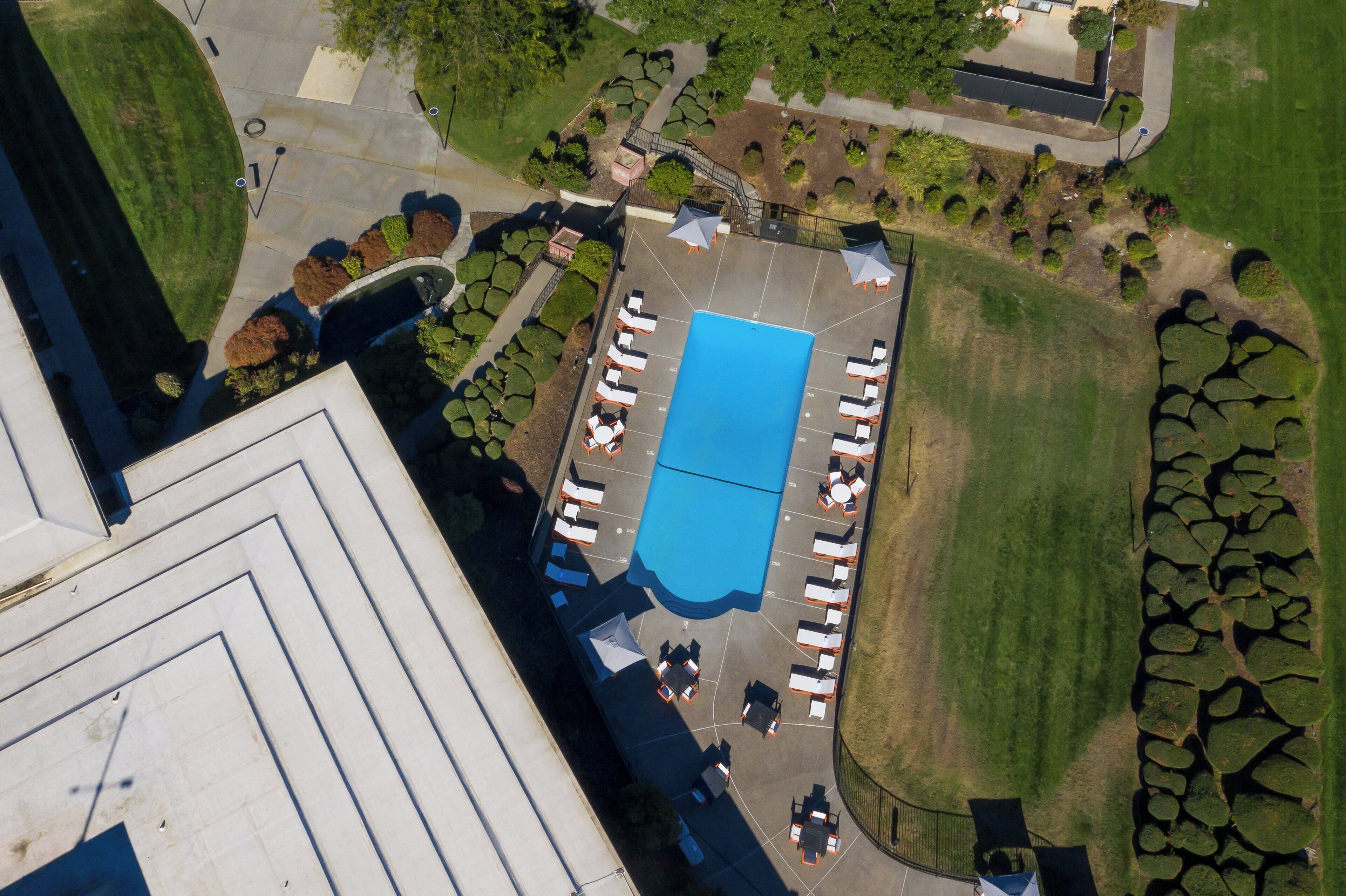 An aerial view of a hotel with a swimming pool surrounded by lounge chairs and umbrellas, a staircase, and landscaped areas.