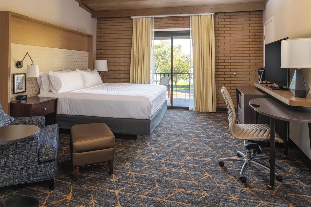 Ein Hotelzimmer mit einem großen Bett, einem Schreibtisch mit Computer, einem Sessel, einem kleinen Tisch und einem Fernseher.