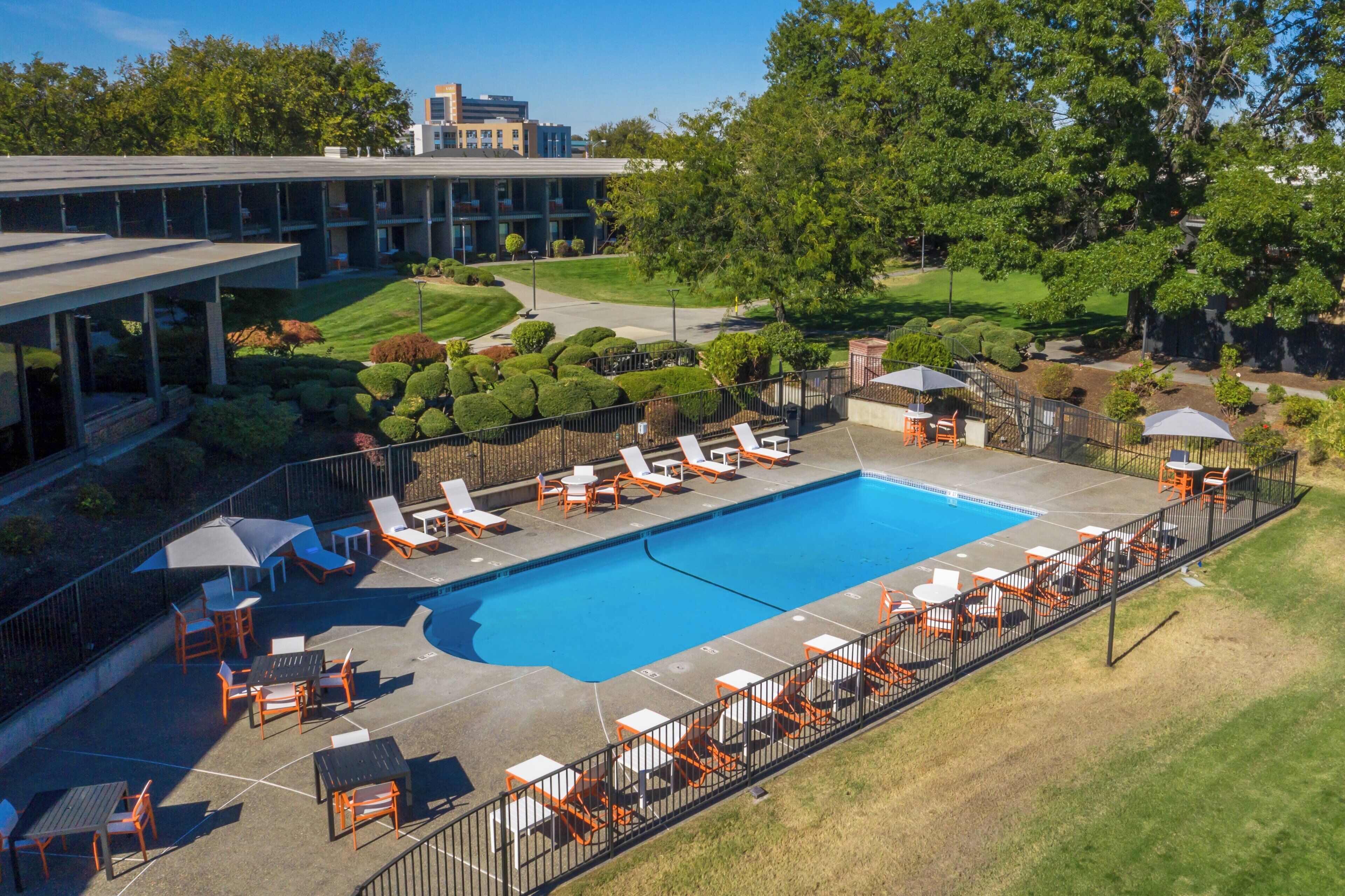 A swimming pool with sun loungers and tables, surrounded by a fence, with a building and trees in the background.