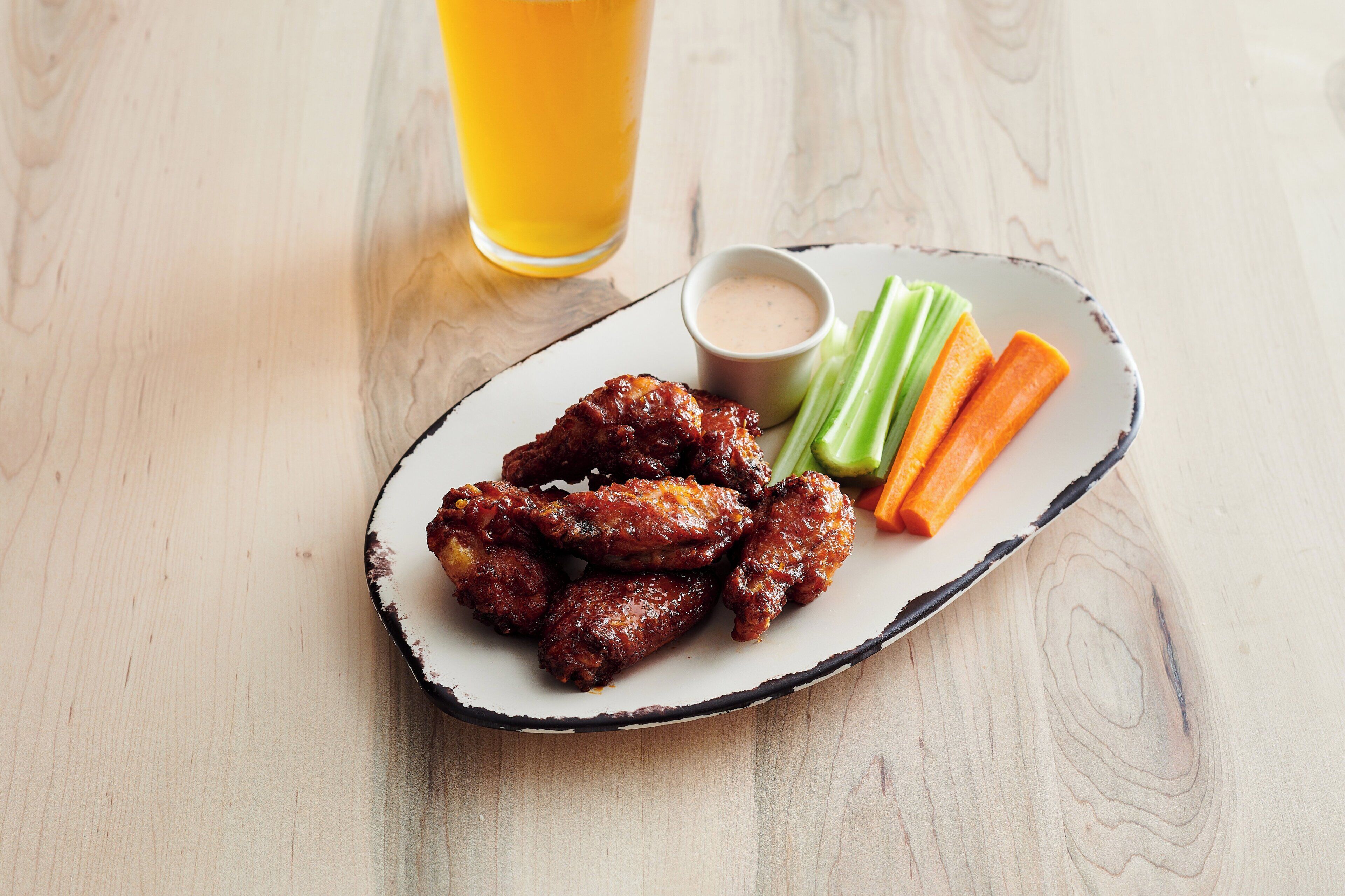 A plate of food with wings, celery, and carrots, and a glass of yellow beverage on a wooden table.