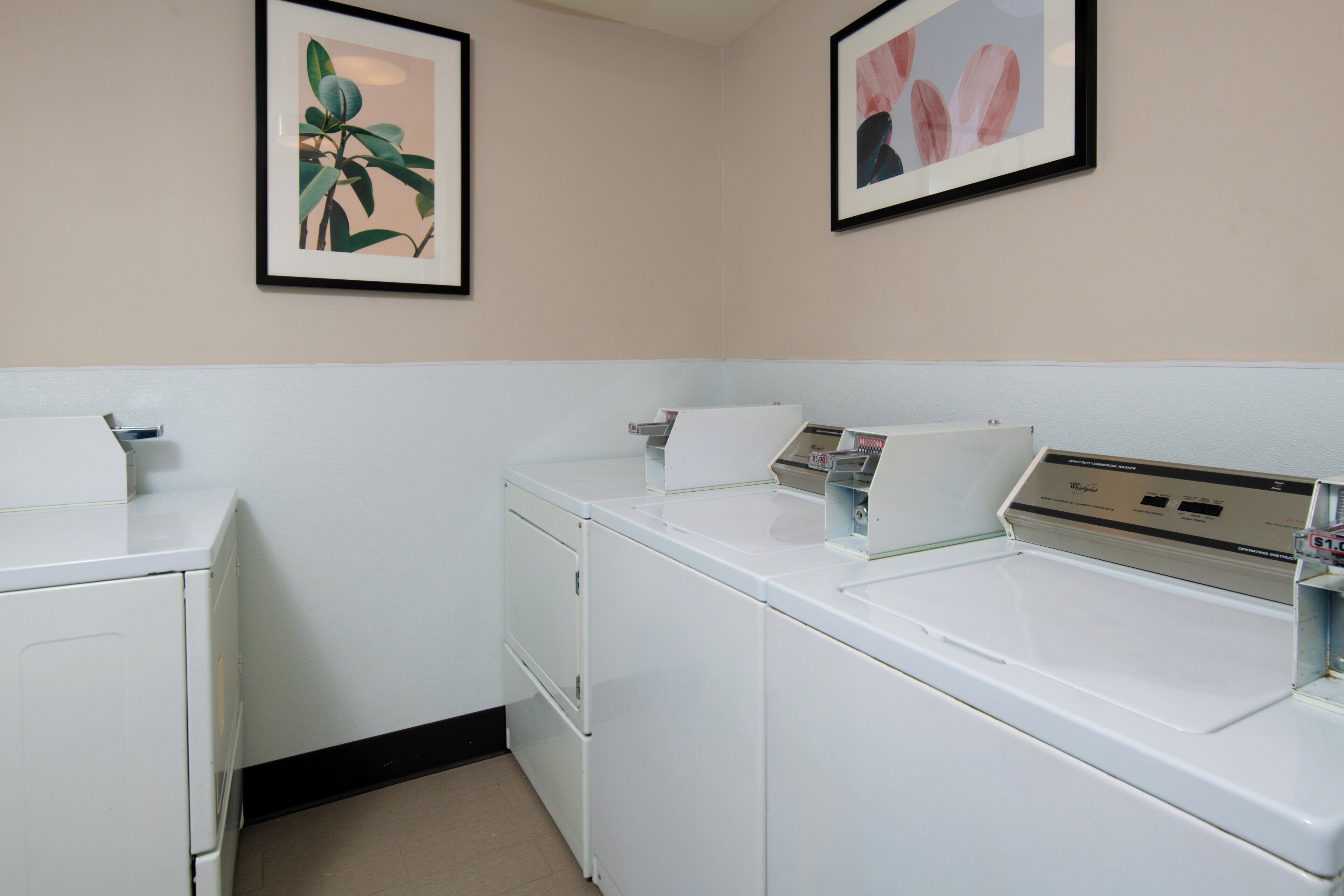 A laundry room with two white washers and dryers, a small shelf with a book, and two framed pictures on the wall.