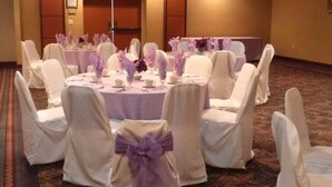 Un salón de banquetes con mesas redondas dispuestas para un evento formal, rodeado de sillas blancas con cintas púrpuras.