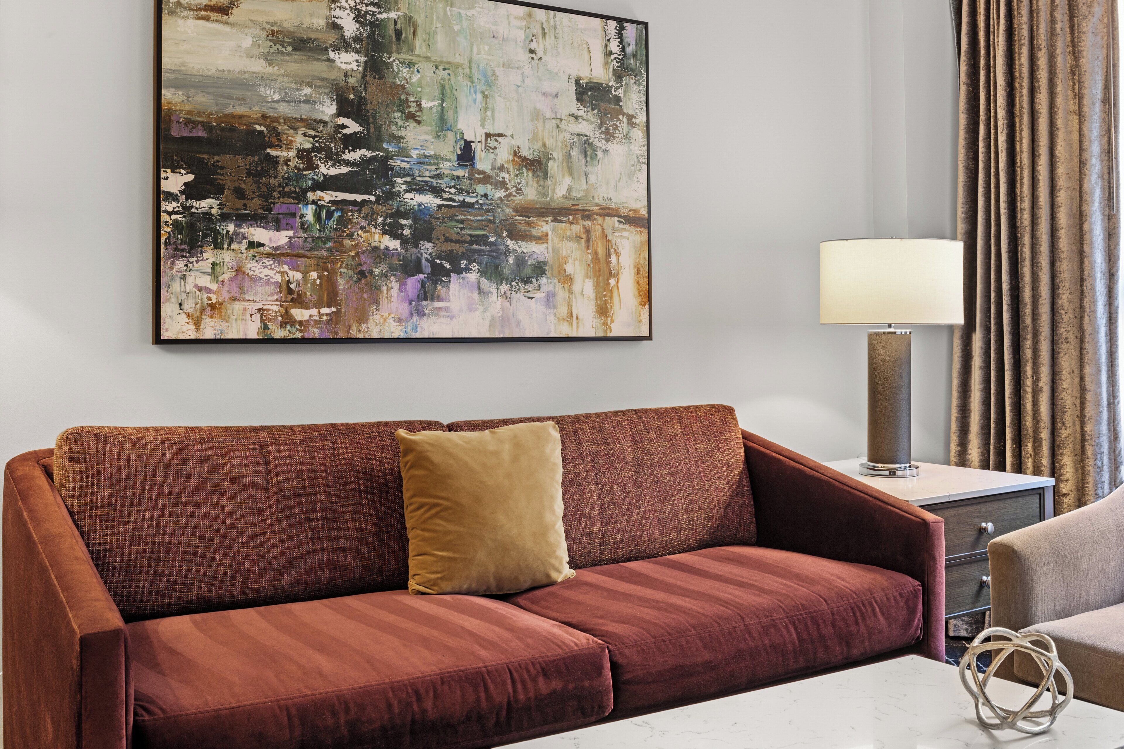 A modern living room with a red sofa, a beige armchair, and a marble coffee table. A large abstract painting is on the wall, and a floor lamp is on a side table.