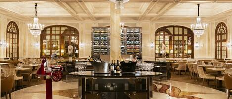 A luxurious hotel lobby with a central bar, chandeliers, and a wine display.