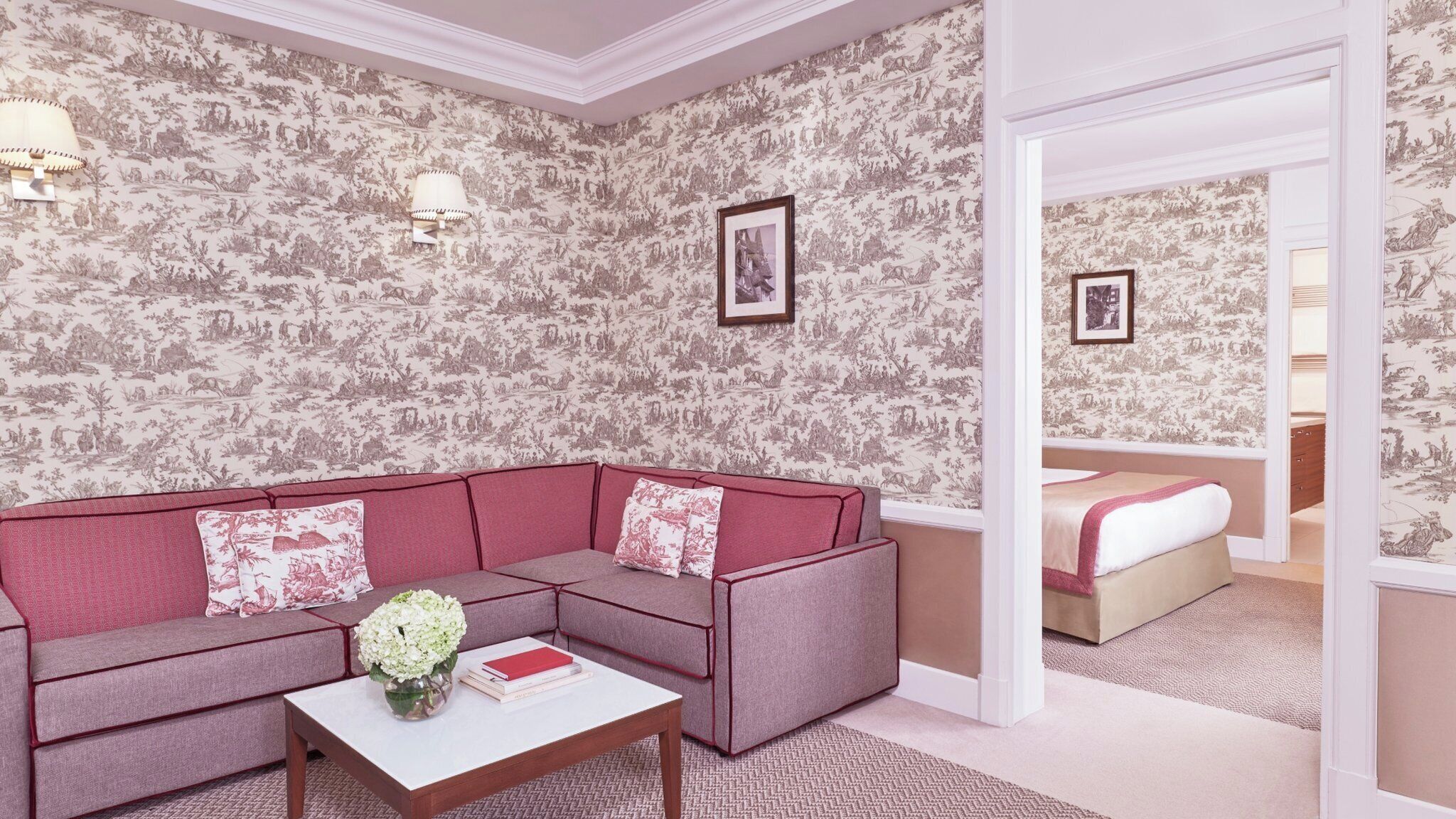 A modern hotel room with a sectional sofa, a small table with a vase and books, and a bed with a pink blanket.