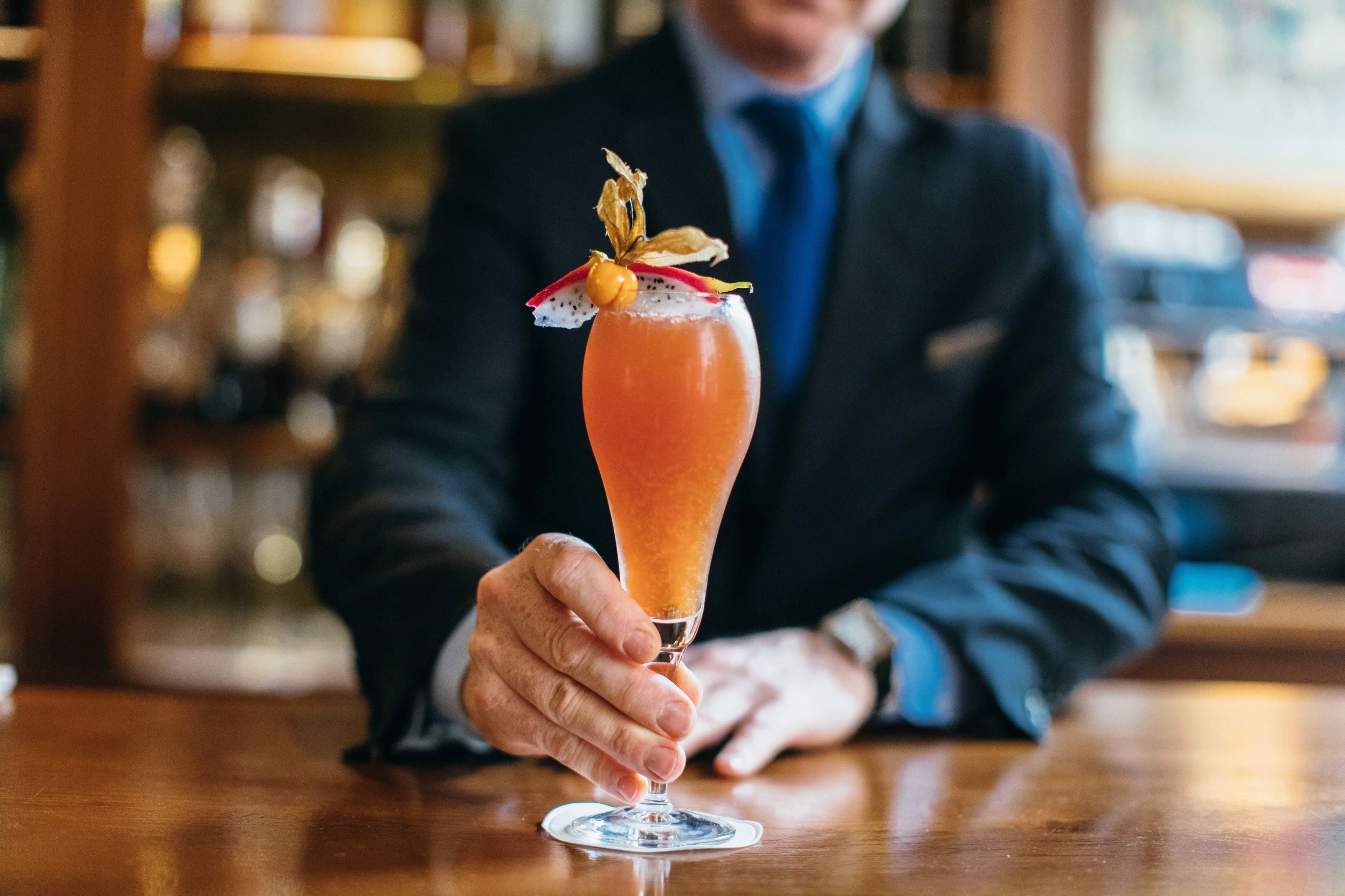 Une personne tenant un verre à cocktail, agrémenté d’une garniture, sur le comptoir en bois d’un bar.