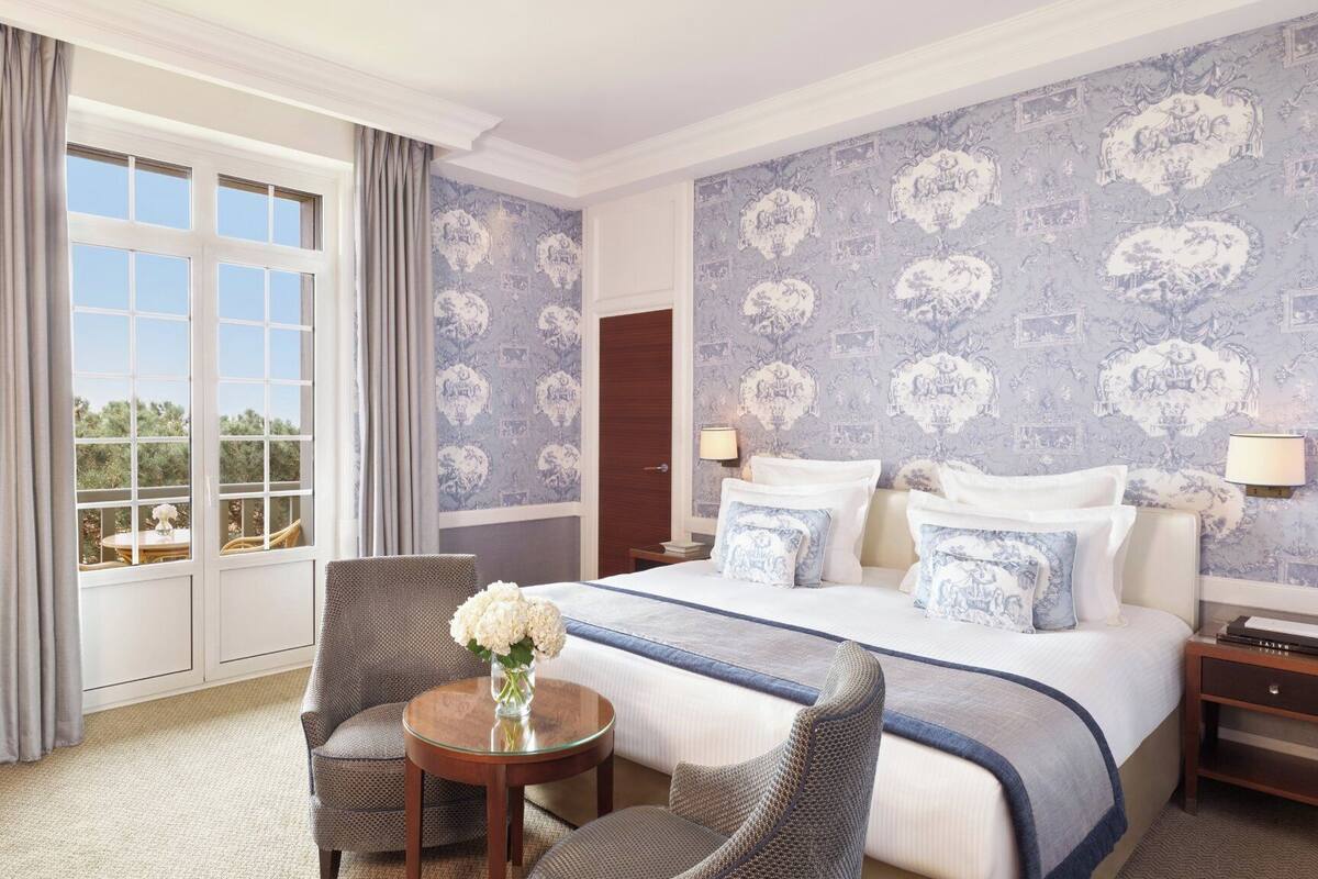 A hotel room with a large bed, two armchairs, a small table with a vase, and a view of greenery through the window.