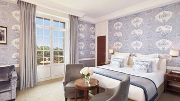 A hotel room with a large bed, two armchairs, a small table with a vase, and a view of greenery through the window.