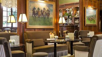 A room with a brown sofa, striped armchairs, a small round table with a bar sign, and a painting of horses on the wall.